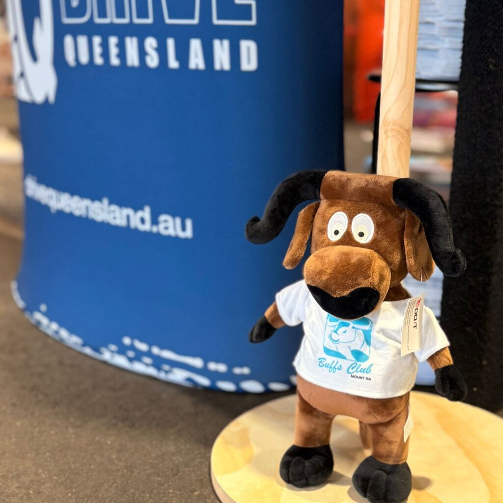 A plush dog toy with curved horns and a white shirt stands on a round wooden base in front of a blue Queensland promotional display.