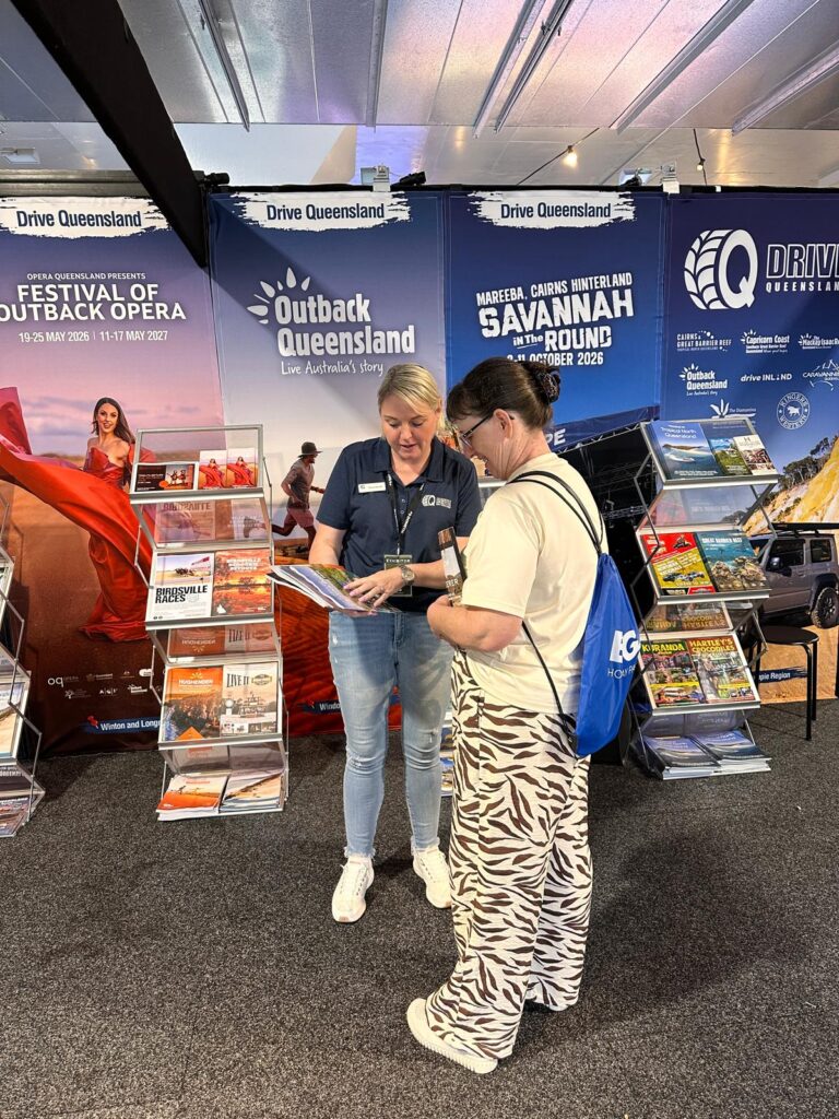 Two women stand at a tourism booth, looking at brochures. Behind them are displays with travel guides and banners for Outback Queensland and Savannah Way events.