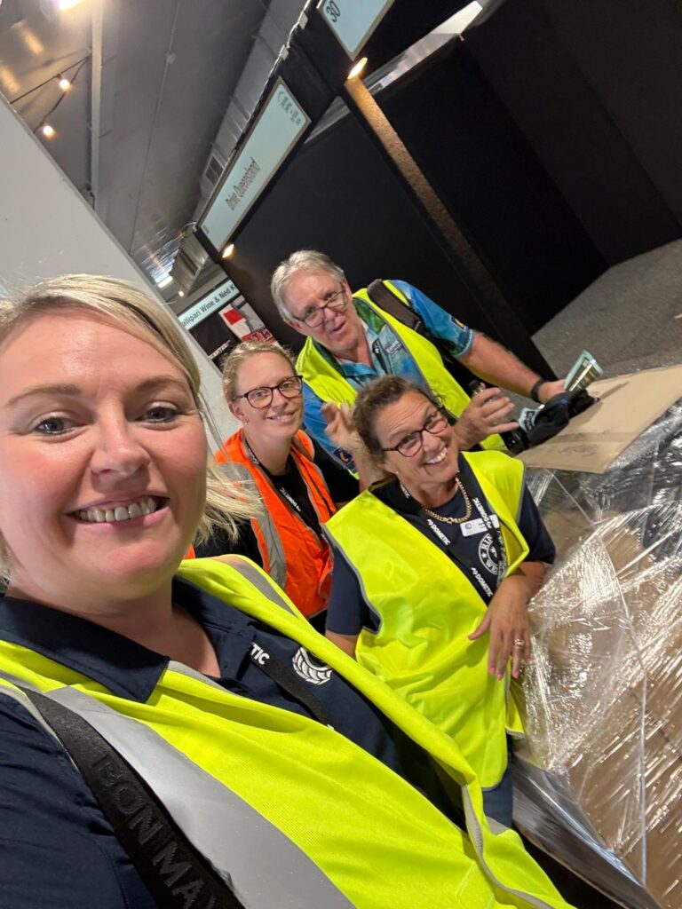 Four people wearing high-visibility vests and glasses smile at the camera while standing near a pallet wrapped in plastic indoors.