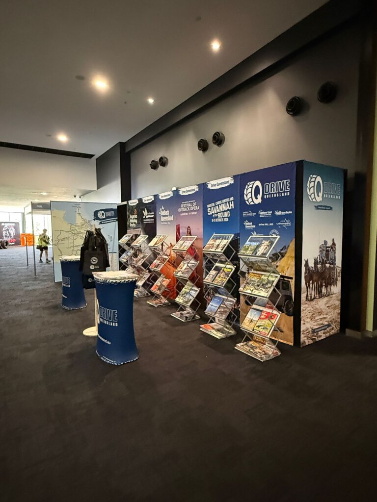 A hallway display features several racks of brochures and magazines promoting tourism, with branded banners and posters lining the wall on a dark carpeted floor.