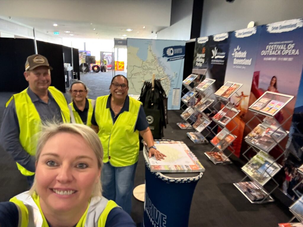 Four people in yellow safety vests stand and smile at an indoor event booth with brochures and promotional materials displayed behind them.