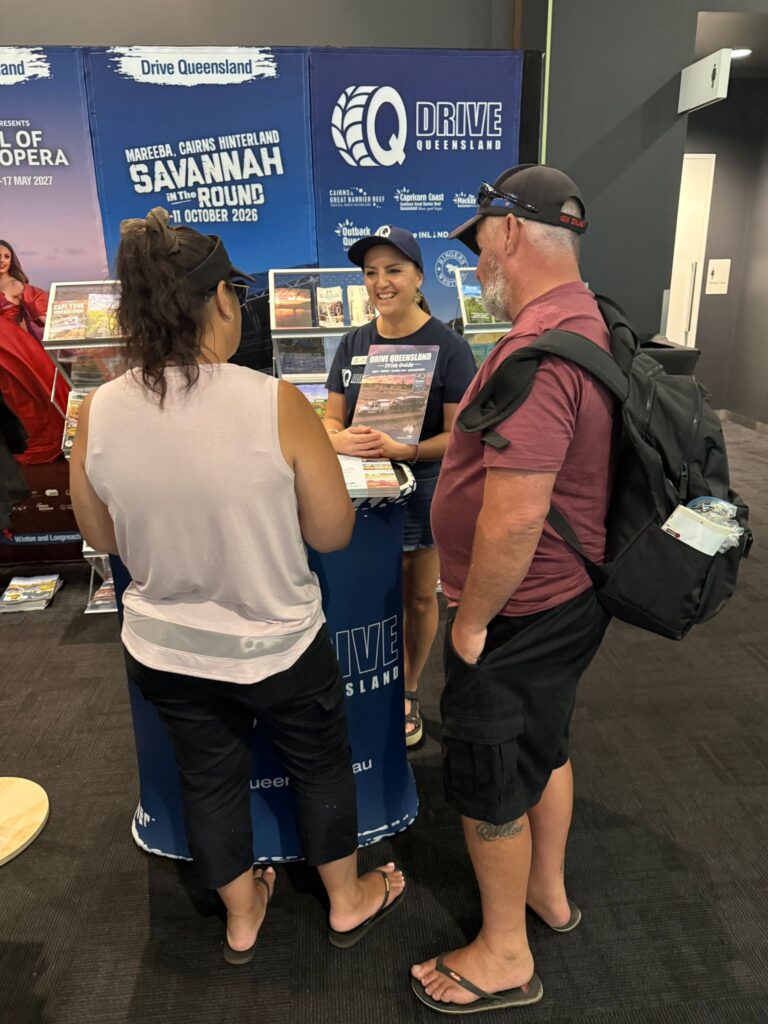 Three people stand at a promotional booth for Drive Queensland, with brochures displayed and posters about Savannah Way and Queensland travel in the background.