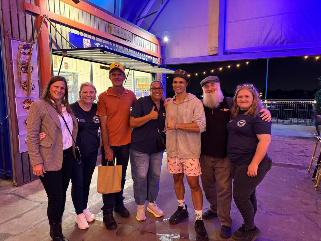 A group of seven people stand together smiling for a photo under blue and purple lights at an outdoor event.