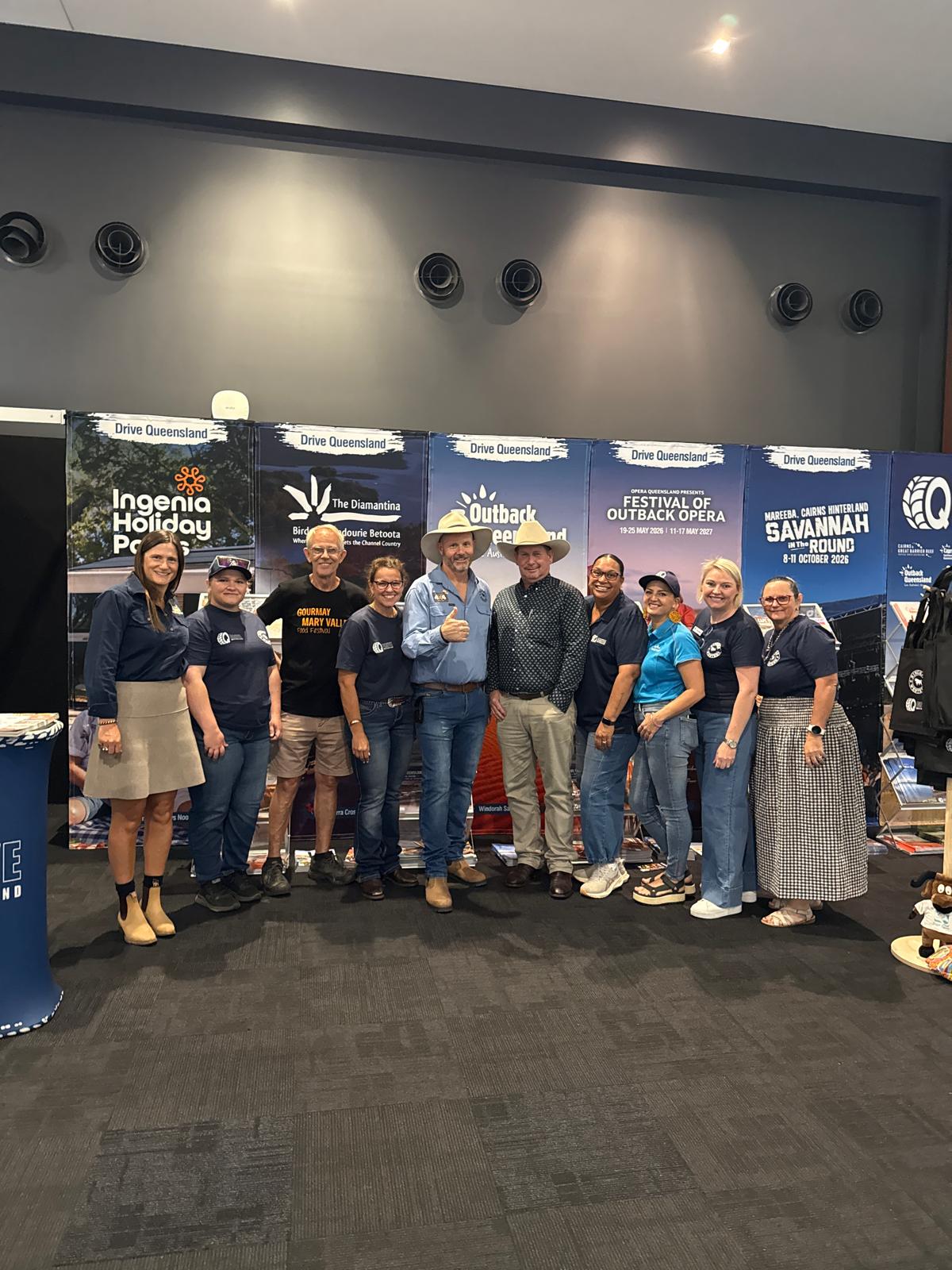 A group of eleven people stand together smiling in front of promotional banners at an indoor event.