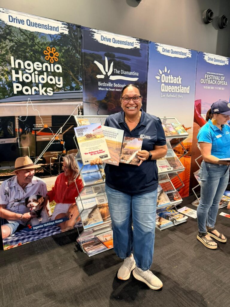 A woman stands in front of tourism display boards for Queensland, holding brochures and smiling. Another person in a blue shirt is visible to the right, arranging pamphlets.