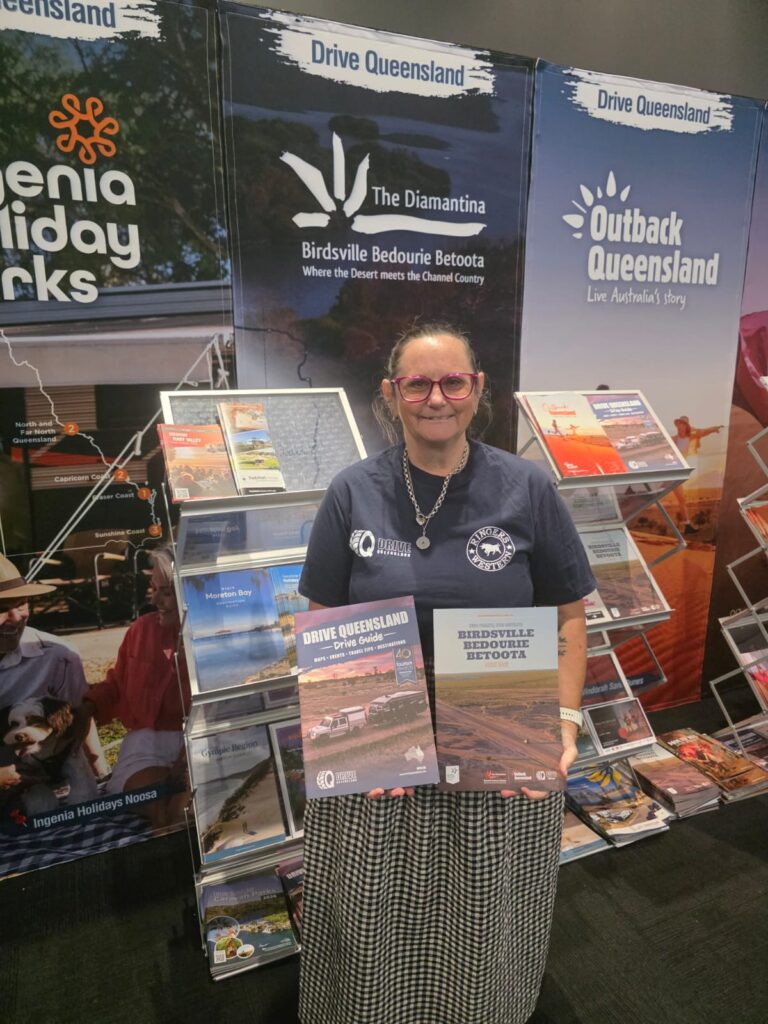 A woman stands indoors holding tourism brochures, in front of a display for Queensland travel information and brochures about Birdsville, Bedourie, and Outback Queensland.