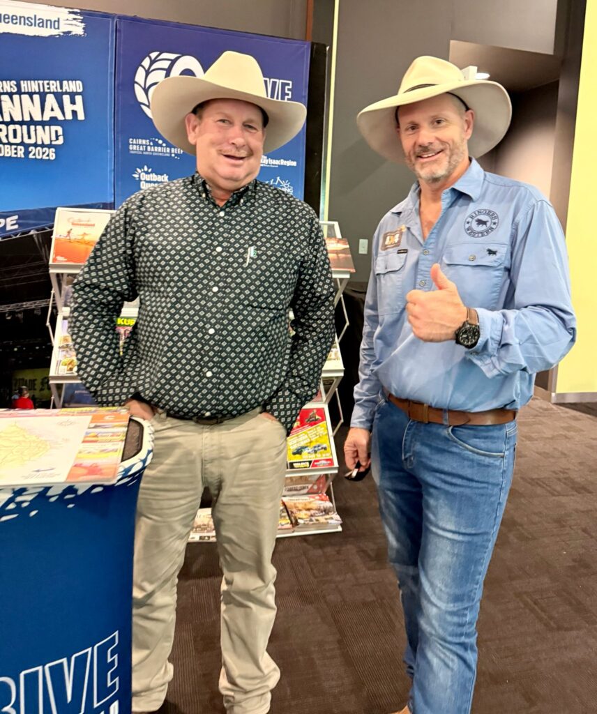 Two men in cowboy hats and casual shirts stand indoors near a promotional booth, one smiling and the other giving a thumbs-up gesture.