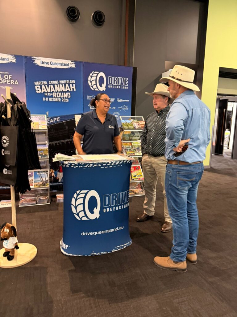 Three people wearing hats stand and talk at a Drive Queensland promotional booth with brochures and banners in the background.