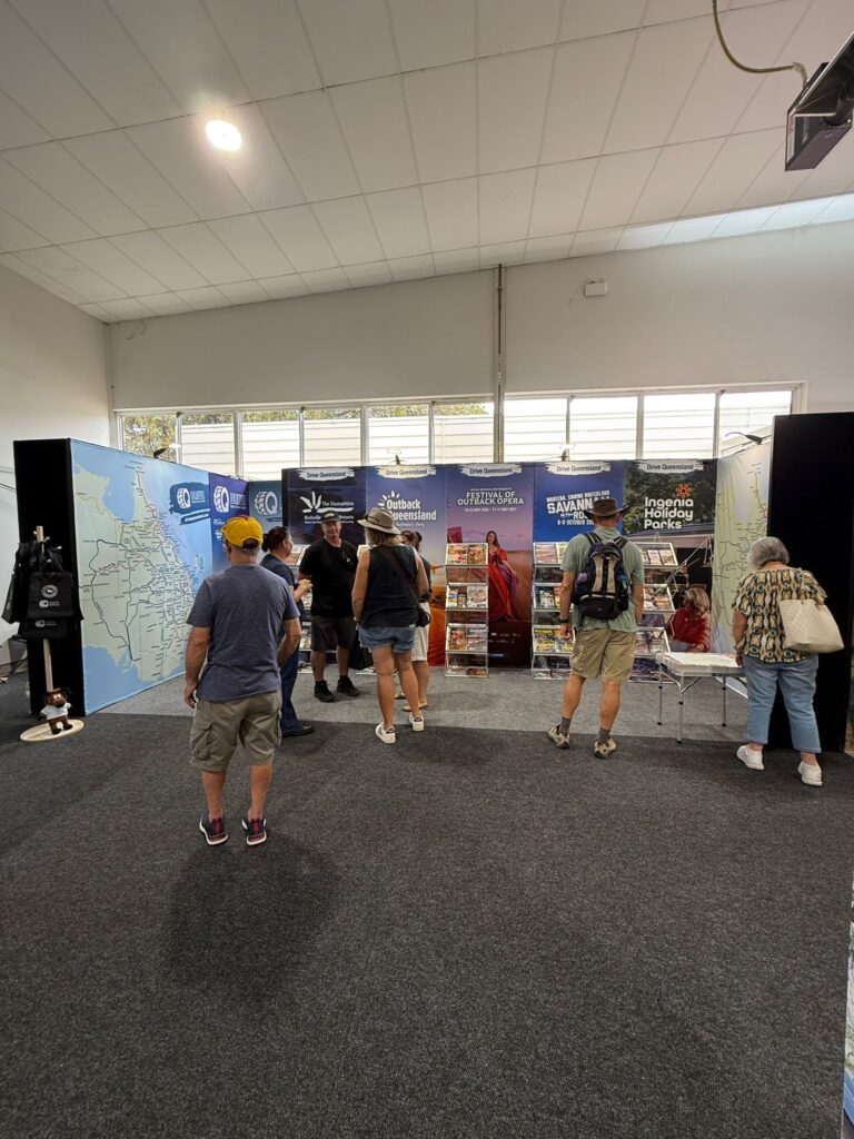 Several people stand and look at tourism display panels and brochures in an indoor exhibition space with carpeted flooring and large windows.