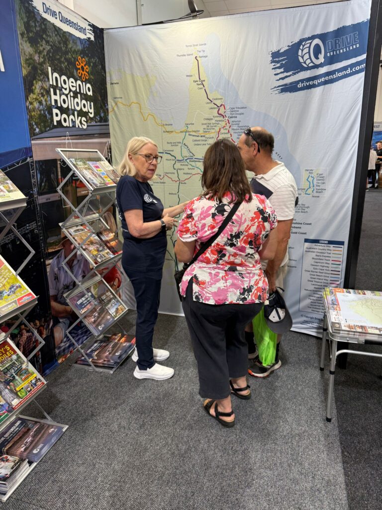 A woman explains a map of Queensland to two visitors at a Drive Queensland booth, with brochures displayed nearby.