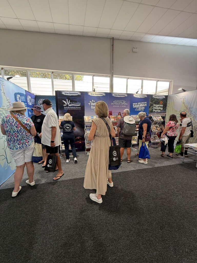 Several people stand in front of promotional booths at an indoor event, reading brochures and carrying tote bags.