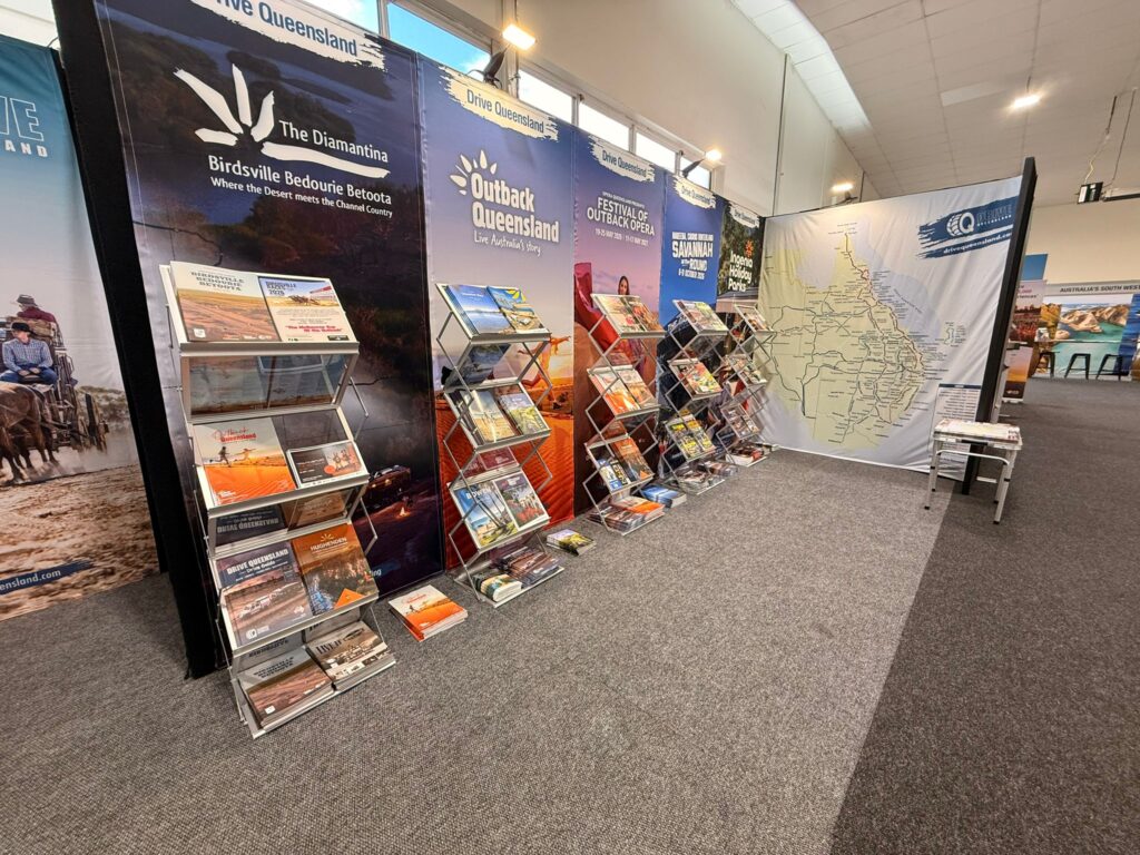 Travel brochures and maps displayed on racks at a tourism information booth, with posters and a large map of Queensland on the wall.