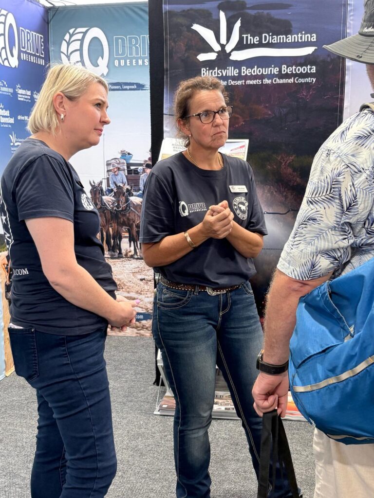 Two women in navy shirts stand talking to a man at an indoor event booth promoting travel in Queensland, with promotional banners in the background.