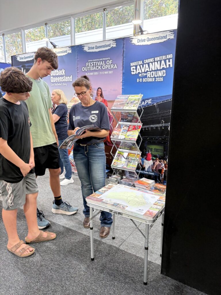 A woman reads a brochure at a display table with tourism flyers, while two young men stand nearby at a travel expo booth promoting Queensland events and destinations.