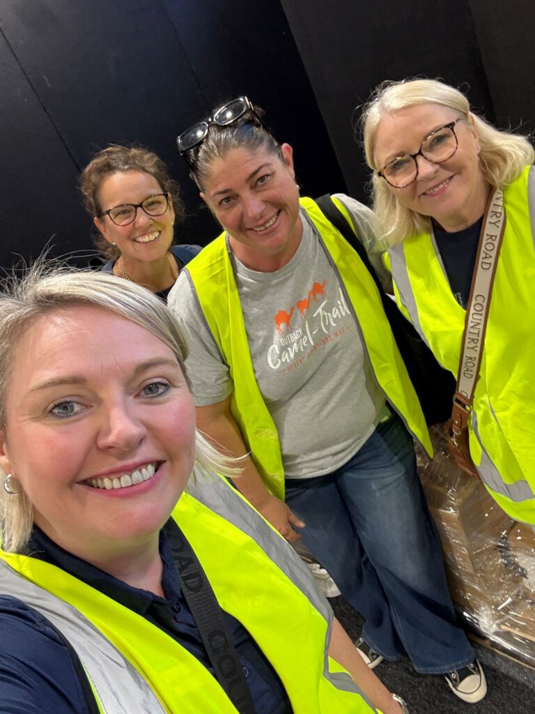 Four women wearing yellow safety vests smile for a selfie indoors. One wears a grey t-shirt with a camel graphic, and another has a beige shoulder bag.