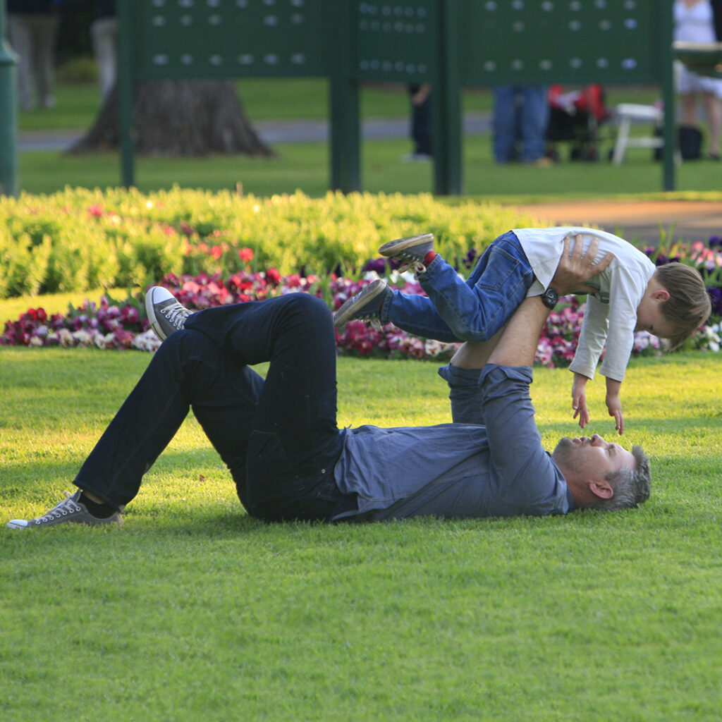 An adult lies on grass in a park, holding a small child up in the air. They are both smiling and surrounded by flower beds and greenery.