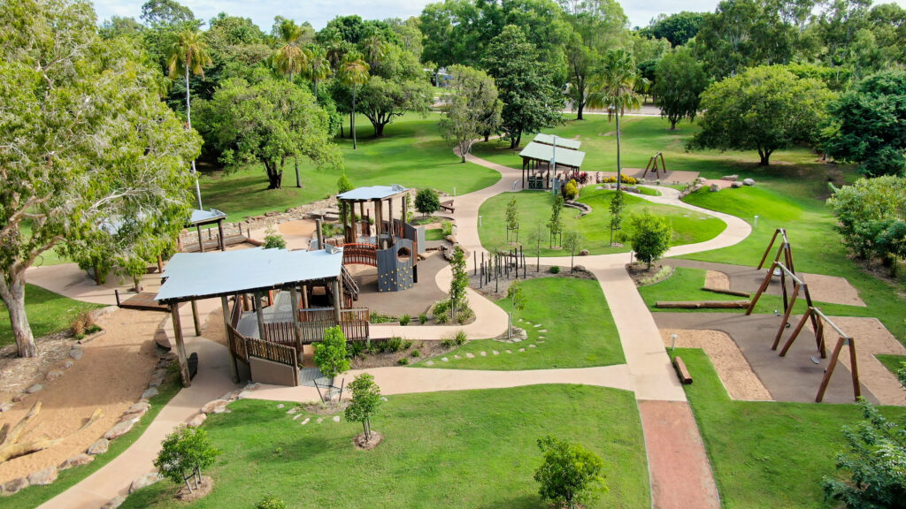 Aerial view of a park with playground structures, swings, covered seating areas, walking paths, and green lawns surrounded by trees.