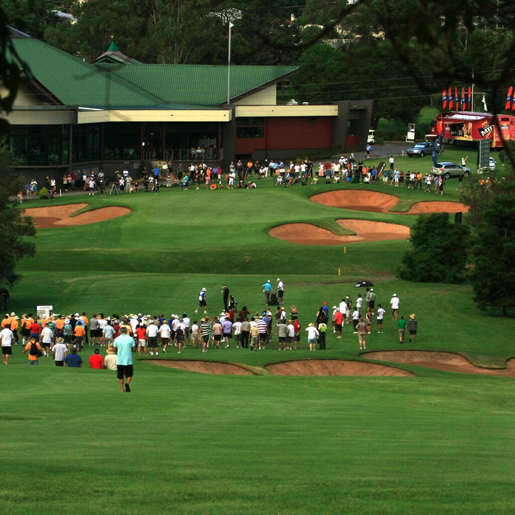Crowd gathered on the fairway of a golf course near a clubhouse, watching golfers play; sand bunkers and trees surround the green.