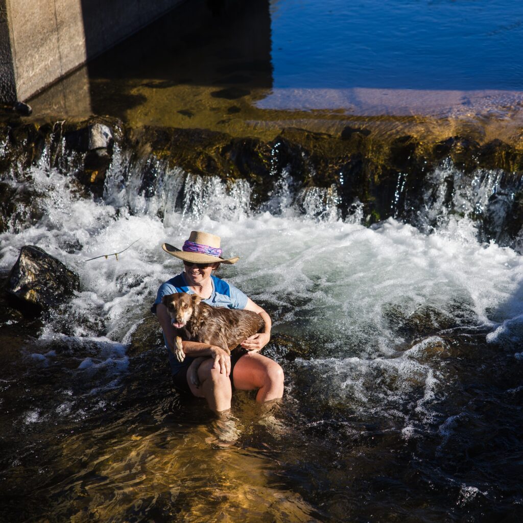 A person wearing a hat sits in shallow water near a small waterfall, holding a brown dog on their lap.