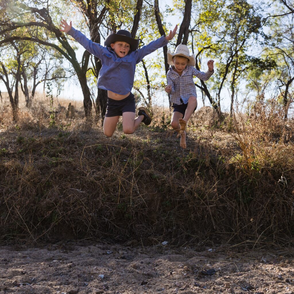 Two children wearing hats jump off a grassy ledge outdoors, with trees and dry grass in the background on a sunny day.