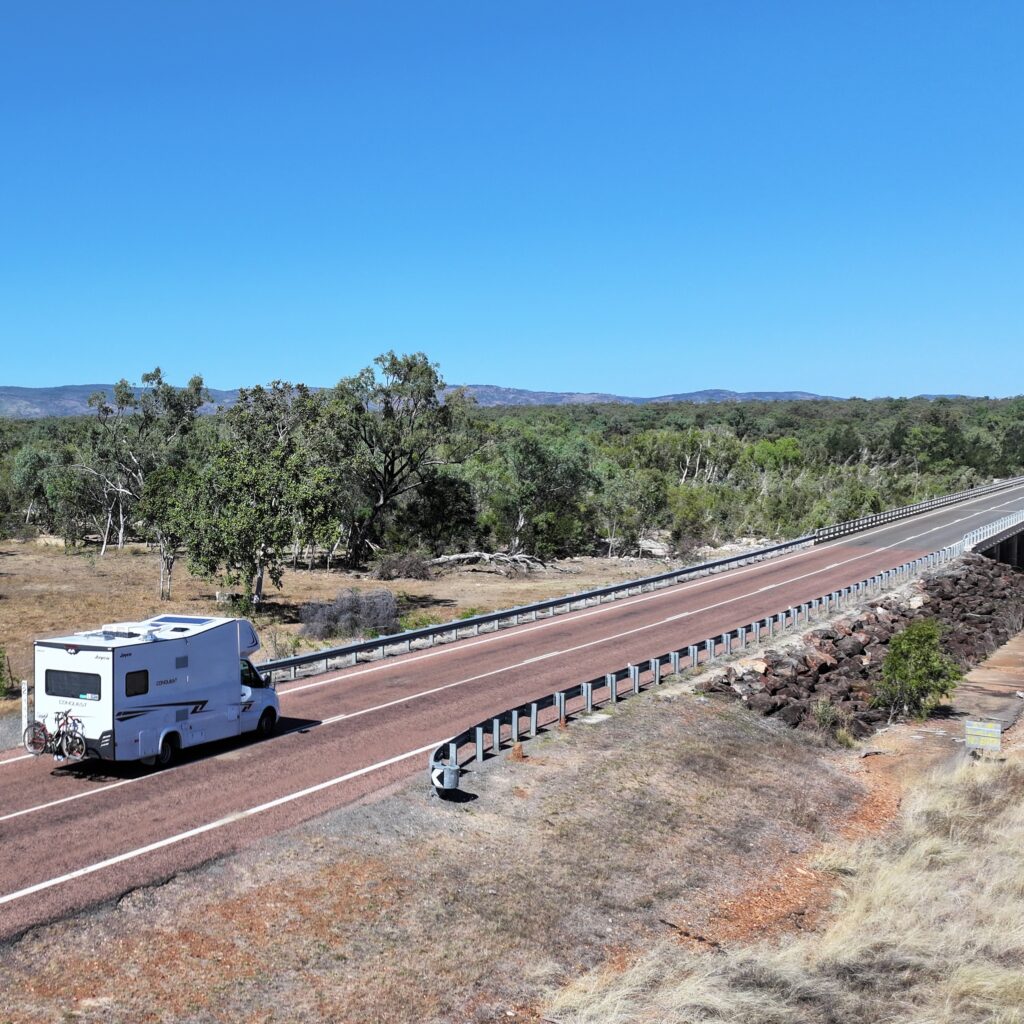 A white camper van drives on a deserted road through a rural landscape with trees and distant hills under a clear blue sky.