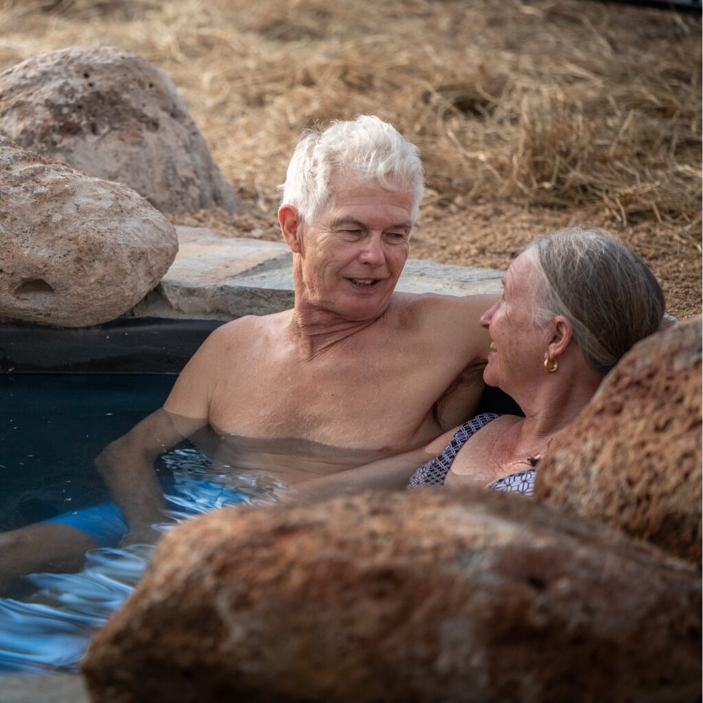An older man and woman relax together in a rocky outdoor hot tub, looking at each other and smiling. Dry grass is visible in the background.