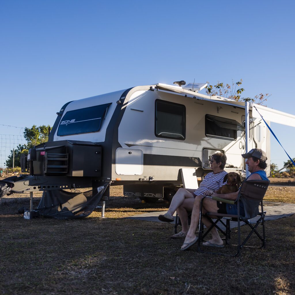 Two people sit on camping chairs with a small dog beside a parked caravan on dry ground under a clear blue sky.