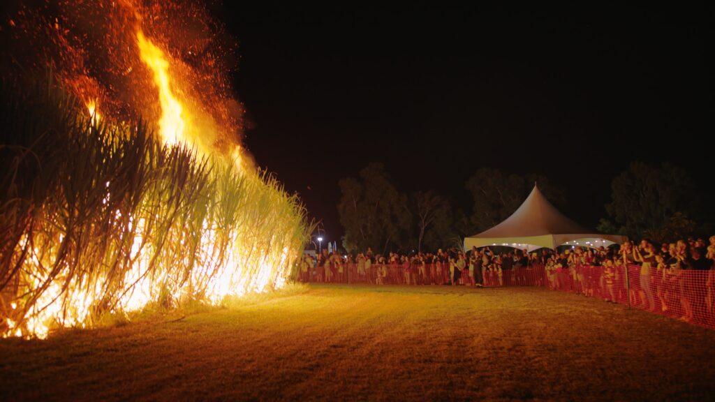 A large fire burns tall grass at night while a crowd watches from behind a barrier near a white event tent.
