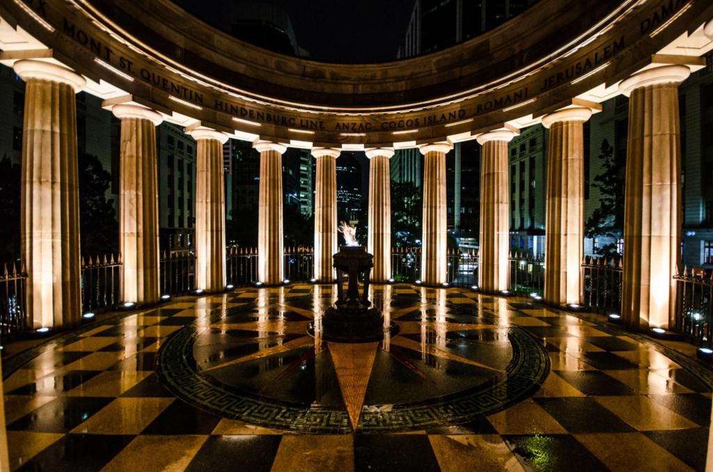 Circular monument with tall columns, an eternal flame in the center, and engraved names along the upper edge, illuminated at night with reflections on the wet checkered floor.
