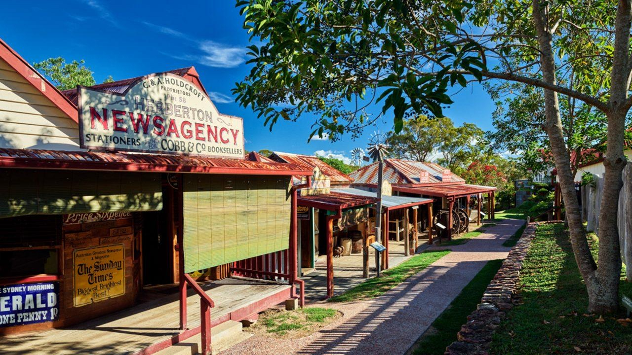 A row of vintage wooden shopfronts, including a newsagency, lines a shaded path under trees on a sunny day.