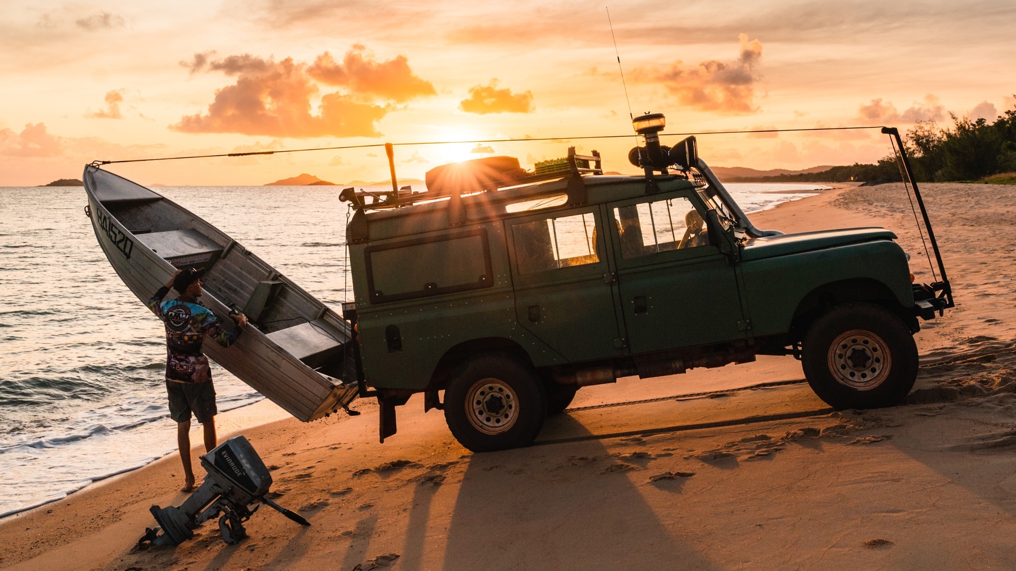 A person loads a small boat onto a green off-road vehicle parked on a sandy beach at sunset, with the sea and distant islands in the background.