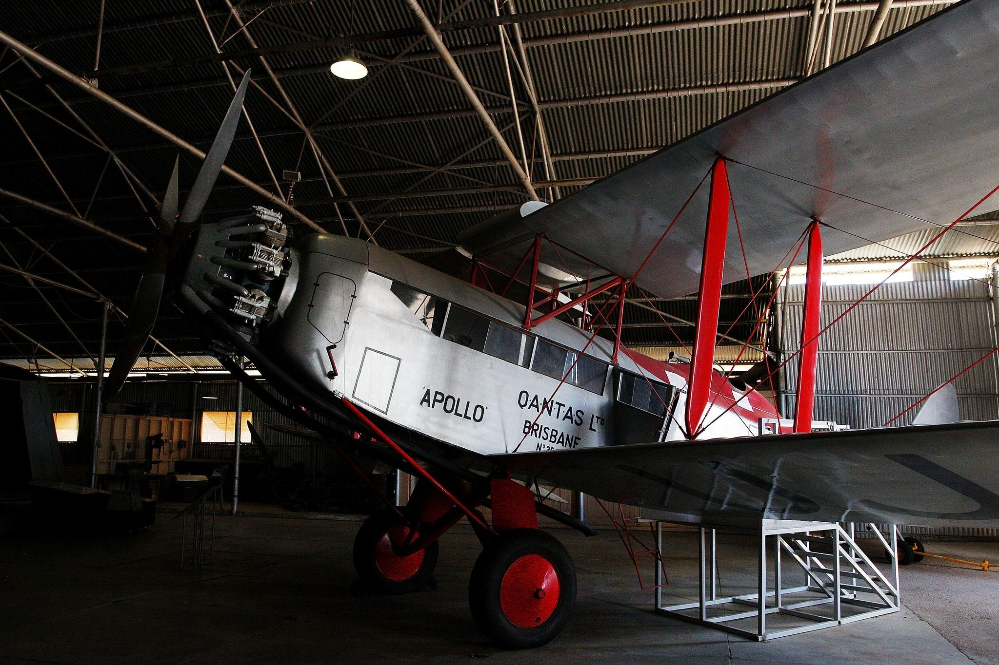 A vintage biplane with "APOLLO QANTAS LTD BRISBANE" written on the side is parked inside an aircraft hangar.