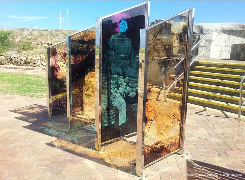 Outdoor art installation with several vertical glass panels, some featuring a portrait and map designs, positioned on a paved area near steps and grassy terrain.