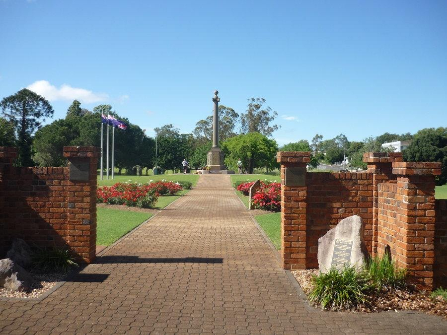 Brick entrance leads to a pathway lined with flowers and flags, leading to a tall memorial monument in a landscaped park under a clear sky.
