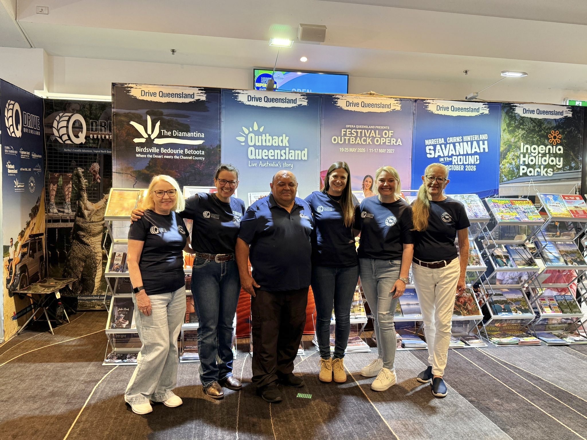 Six people stand side by side in front of tourism display banners and brochures at an indoor event, all wearing matching navy shirts and smiling at the camera.
